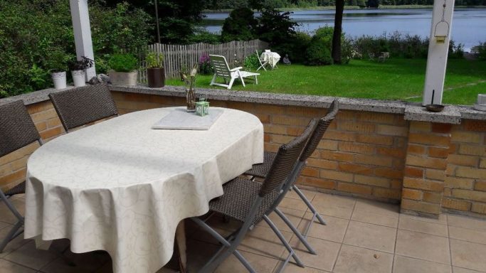 mit einer weißen Tischdecke belegter Tisch mit vier Stühlen und Blick über die Wiese in Richtung See